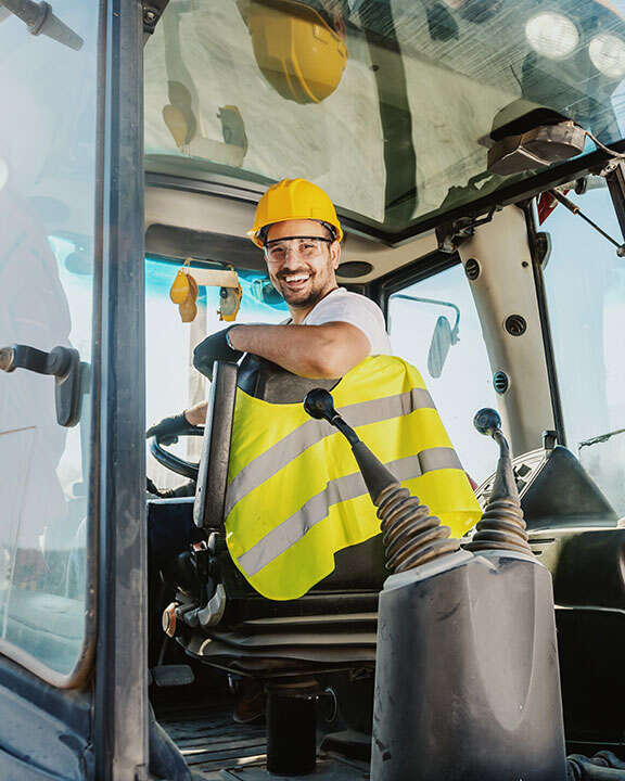 A construction worker driving an excavator backwards as they are looking to towards the camera